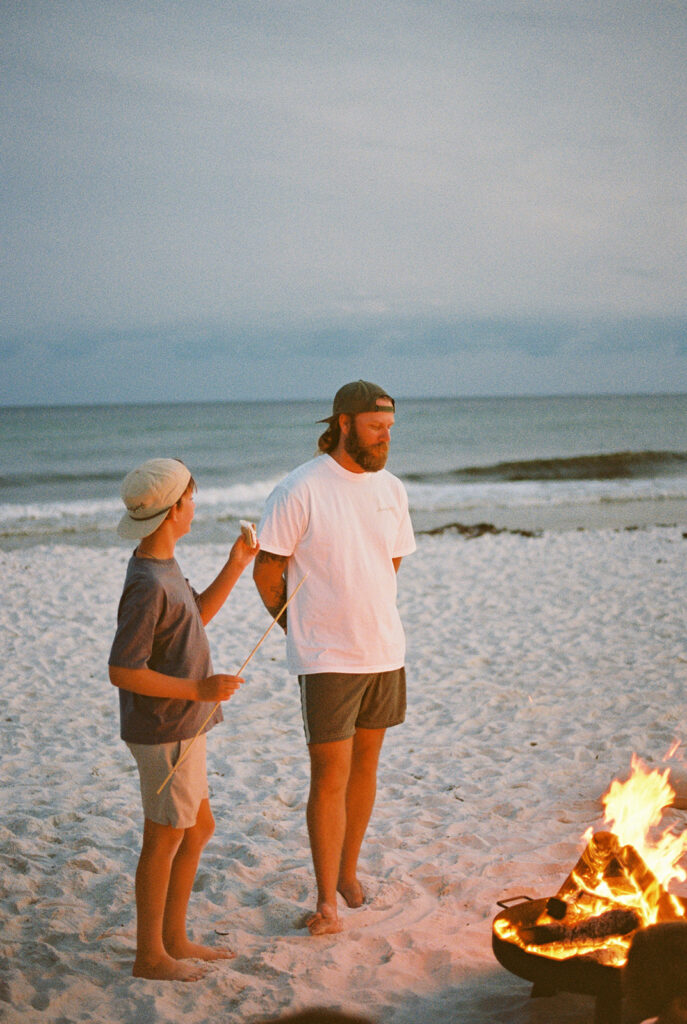 A husband and son around the fire putting a marshmallow on a stick to make a smore. The fire is lit around them.